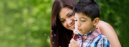 A happy mom watching her young son blowing on a dandelion.